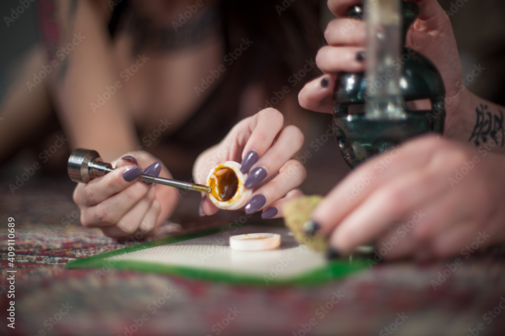 Two young ladies use a metal dabber to scoop concentrated THC cannabis wax, shatter, oil out of silicone container to smoke in a chilled custom blown glass bong with ice cubes Two young ladies use a metal dabber to scoop concentrated THC cannabis wax, shatter, oil out of silicone container to smoke in a chilled custom blown glass bong with ice cubes
