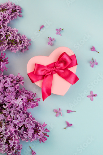 Heart shape pink gift box with red ribbon bow, purple Lilac branch flower blooming bouquet on blue background. Summer Valentine's day love creative composition. Beautiful greenery blossom © Julia Klintsova