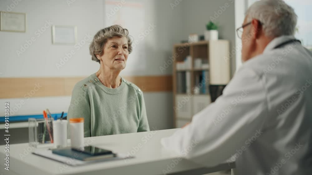 Middle Aged Family Doctor is Talking with Senior Female Patient During Consultation in a Health Clinic. Experienced Caucasian Physician in Lab Coat Sitting Behind a Computer Desk in Hospital Office. Middle Aged Family Doctor is Talking with Senior Female Patient During Consultation in a Health Clinic. Experienced Caucasian Physician in Lab Coat Sitting Behind a Computer Desk in Hospital Office.