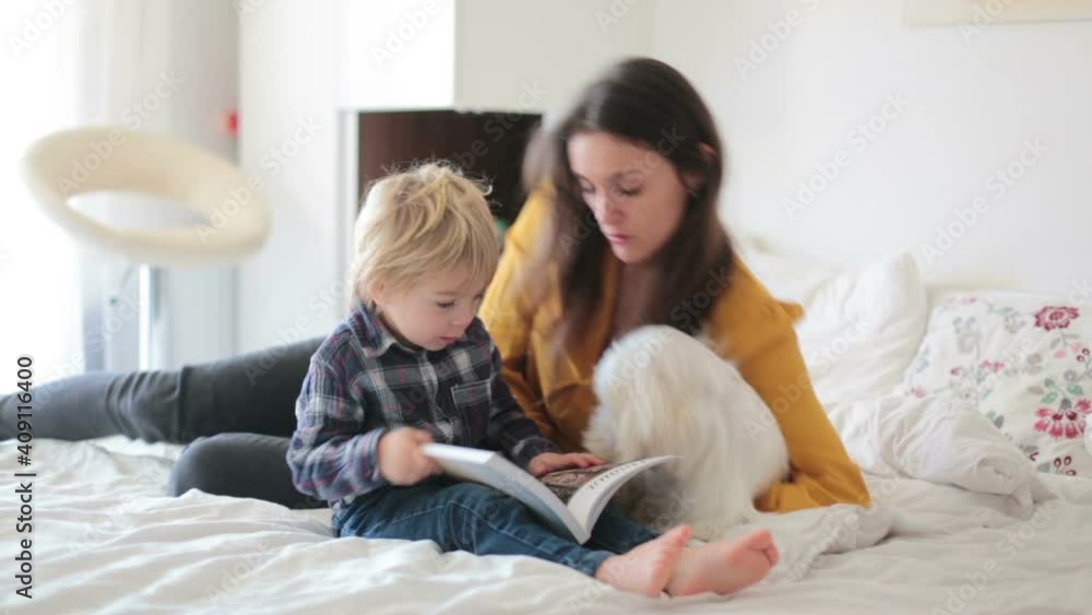 Mother, toddler child and puppy dog, lying on the bed, reading book together, happy family time Mother, toddler child and puppy dog, lying on the bed, reading book together, happy family time