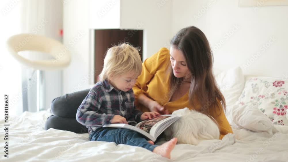 Mother, toddler child and puppy dog, lying on the bed, reading book together, happy family time Mother, toddler child and puppy dog, lying on the bed, reading book together, happy family time