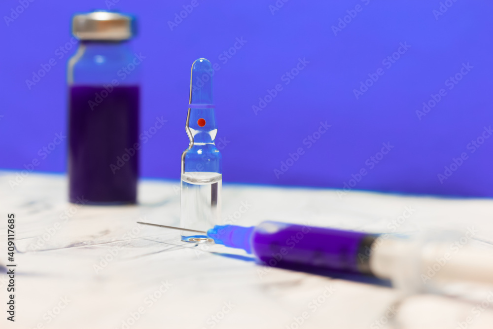 A glass medical transparent ampoule stands on a table on purple wall background. Flu vaccine and syringe isolated, pandemic coronavirus and medical care A glass medical transparent ampoule stands on a table on purple wall background. Flu vaccine and syringe isolated, pandemic coronavirus and medical care