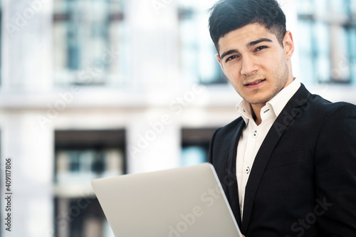 a dark-haired businessman of European appearance stands in a business center in a stylish black suit. holds a laptop computer in his hands. copy space. banner for the site.