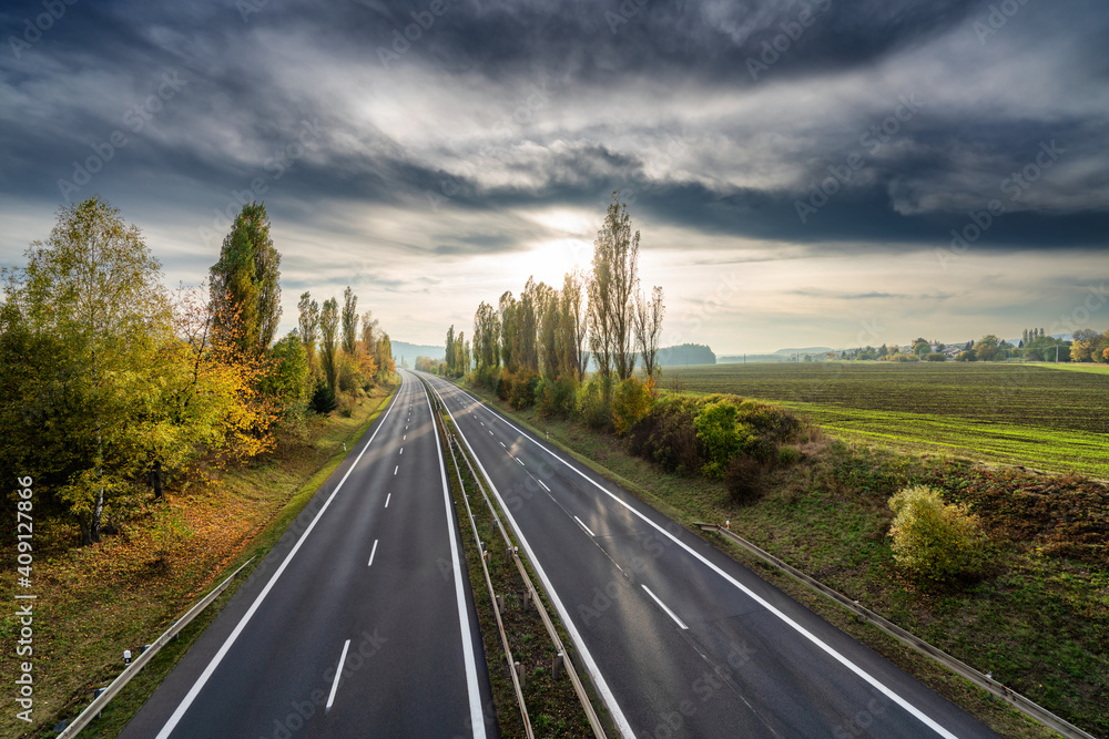 Empty asphalt highway between the avenue of poplars in autumn rural landscape at sunset with dark clouds