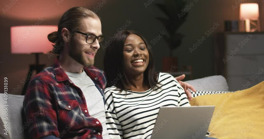 Close up portrait of cheerful young couple of Caucasian man and African American woman speaking and laughing sitting on sofa in house surfing internet, videochatting on laptop computer, love concept Close up portrait of cheerful young couple of Caucasian man and African American woman speaking and laughing sitting on sofa in house surfing internet, videochatting on laptop computer, love concept