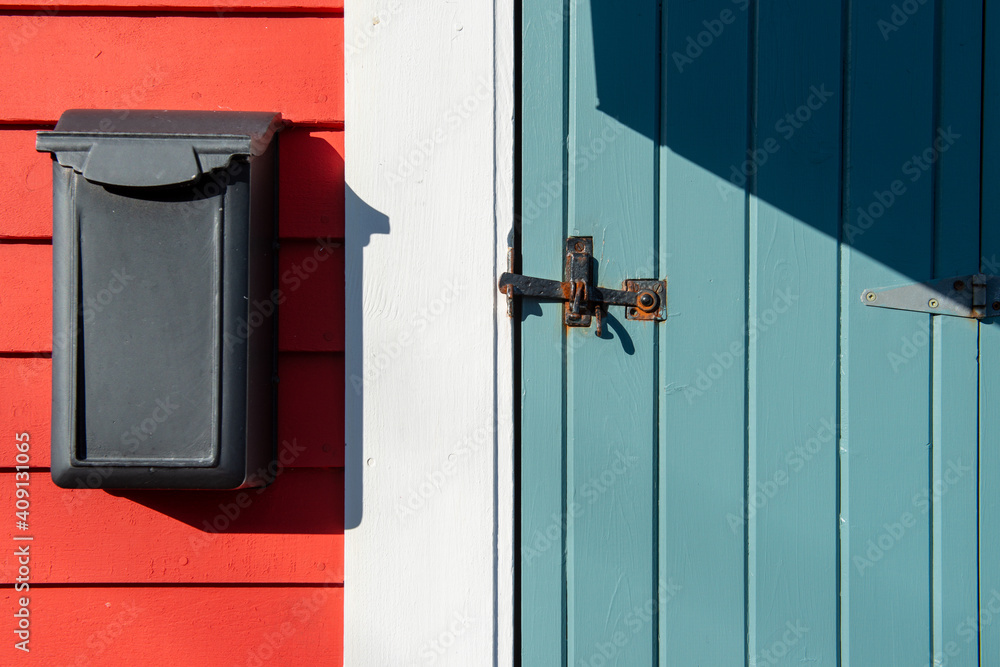 A pale blue wooden strap storm door with antique hinges and a black metal latch. The door is on the exterior of a red house with a long black mailbox or postal box. The door has white trim around it.
