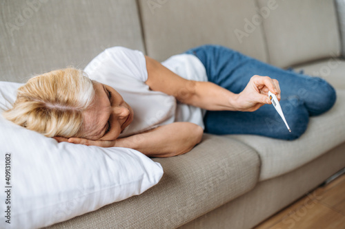 Unhealthy mature woman lying on sofa with headache looks at the thermometer, she has a high fever, virus, cold, illness