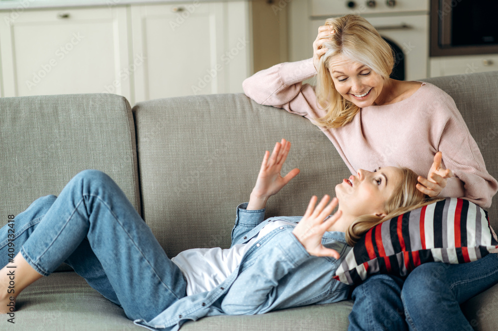 Middle aged attractive caucasian mother and her beloved young adult daughter spend time together on the couch in the living room, chat, gossip, confess, mom gives advice to her daughter Middle aged attractive caucasian mother and her beloved young adult daughter spend time together on the couch in the living room, chat, gossip, confess, mom gives advice to her daughter