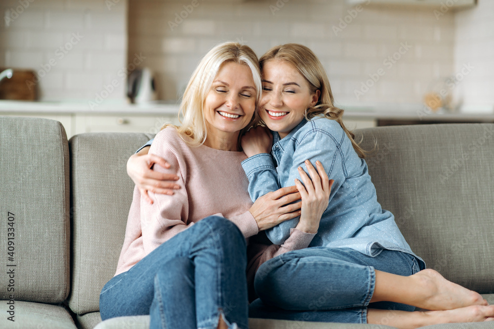 Middle aged blonde caucasian mother and her young adult happy daughter, are sitting at home on the couch at living room, embracing, smiling. Love of mom and daughter