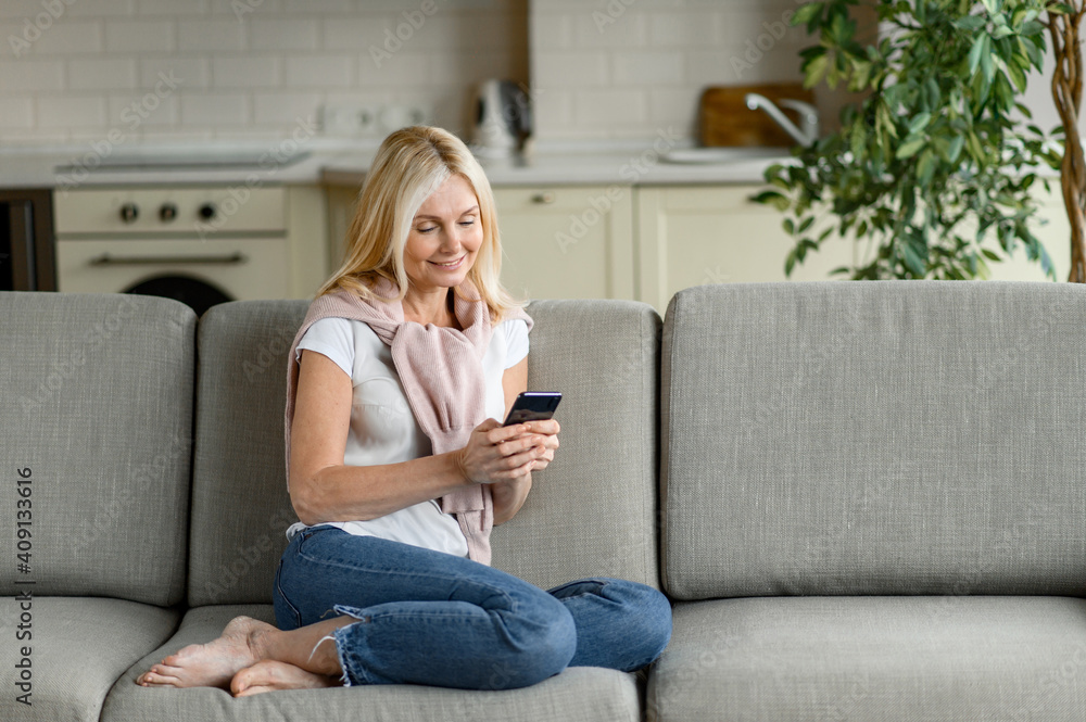 Happy middle aged blonde caucasian woman sits at the sofa, using smartphone. Smiling woman in casual wear chat with friends or family, browsing social network, smiling Happy middle aged blonde caucasian woman sits at the sofa, using smartphone. Smiling woman in casual wear chat with friends or family, browsing social network, smiling