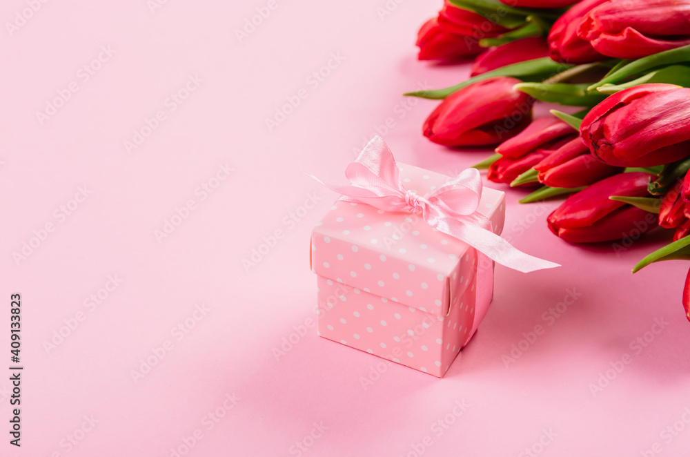 Gift box with a pink ribbon in white polka dots on a background of a bouquet of red tulips with copy space for text. Wallpaper or banner for gift shop, flower shop or jewelry store. Valentine day Gift box with a pink ribbon in white polka dots on a background of a bouquet of red tulips with copy space for text. Wallpaper or banner for gift shop, flower shop or jewelry store. Valentine day