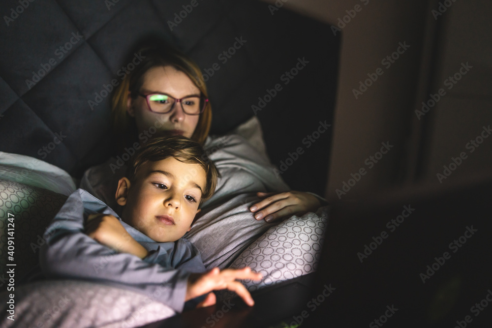 Mother and son watching movie or cartoon using laptop in bed at nighttime - Small boy and his mom using computer while lying in bed at night in dark room - leisure activity family bedtime concept Mother and son watching movie or cartoon using laptop in bed at nighttime - Small boy and his mom using computer while lying in bed at night in dark room - leisure activity family bedtime concept
