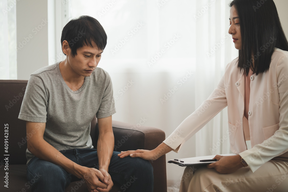 Young asian man suffers from a mental who needs to therapy with a psychologist while sitting on couch to consult, psychiatrist has encouragement the patient by touching to make his feel relaxed. Young asian man suffers from a mental who needs to therapy with a psychologist while sitting on couch to consult, psychiatrist has encouragement the patient by touching to make his feel relaxed.
