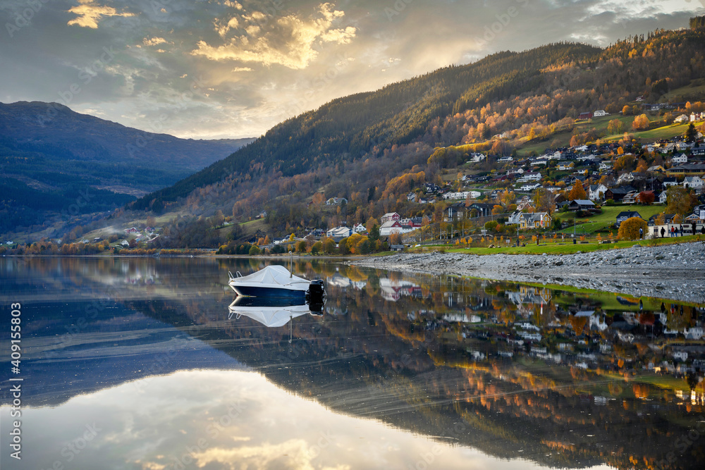 A boat floats in a lake in Voss, Norway, the still water looks like a mirror, reflecting the house on the hill with the trees in the autumn in the evening, the atmosphere is calm and relaxed.