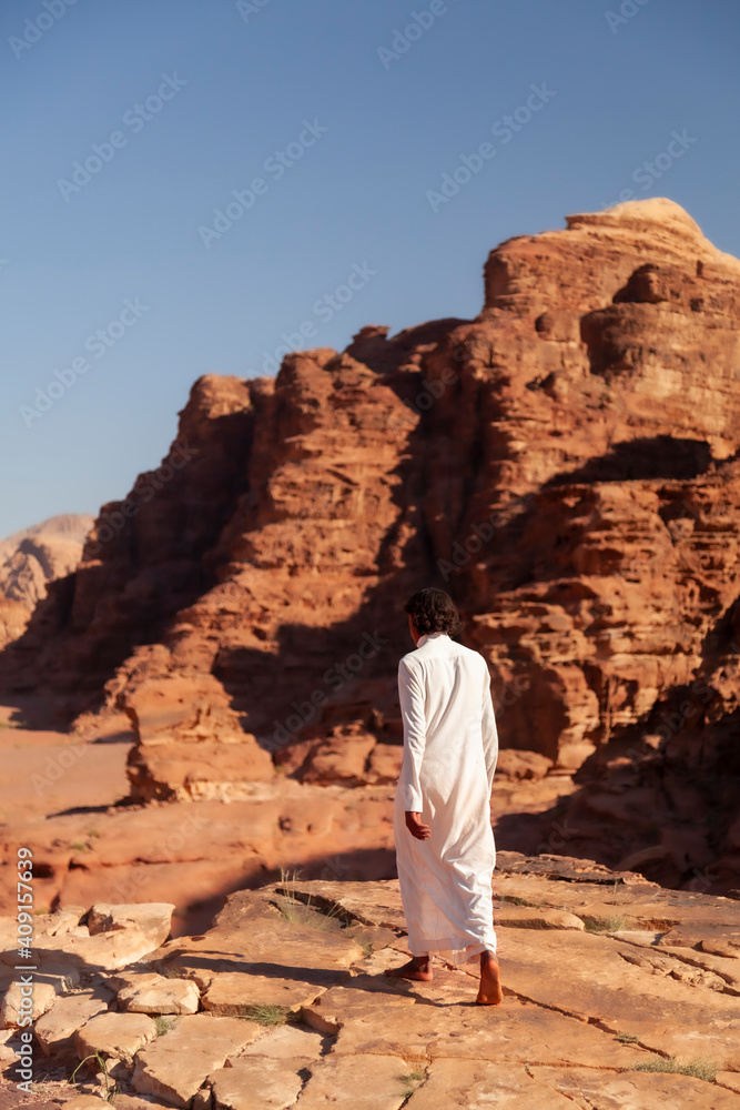 A young Jordanian beduin man wearing white Jubba thobe dress is walking bare feet on top of the cliffs overlooking the Wadi Rum desert. Red sandstone rock formations are seen blurred in  background.