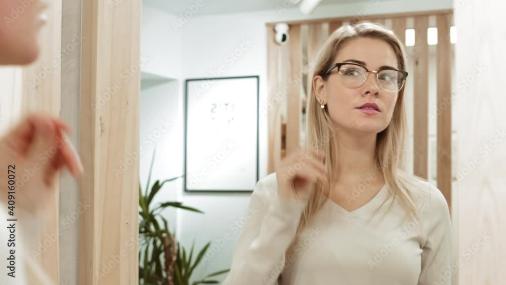Back view of young blond-haired woman standing in front of mirror in optician store, choosing new glasses and then looking at camera with smile on her face Back view of young blond-haired woman standing in front of mirror in optician store, choosing new glasses and then looking at camera with smile on her face