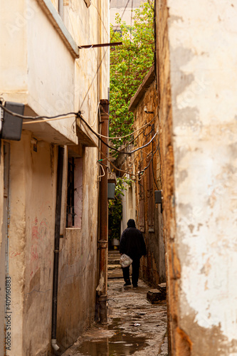 A narrow alley in a low income old city neighborhood in Damascus, Syria. Houses are run down with paint chips cables hanging between buildings. Rain puddles on the street. A man is walking at distance © Grandbrothers