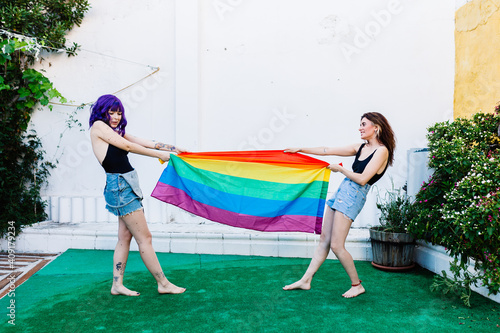 Two women with rainbow flag on a backyard. Couple Young lesbian girls holding a rainbow flag and hugging happily. A pair of lesbian women