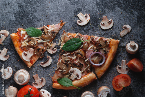 Slices of homemade Italian pizza with mushrooms, ham, tomatoes, cheese, onions and herbs, on a dark slate stone table