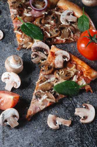 Slices of homemade Italian pizza with mushrooms, ham, tomatoes, cheese, onions and herbs, on a dark slate stone table