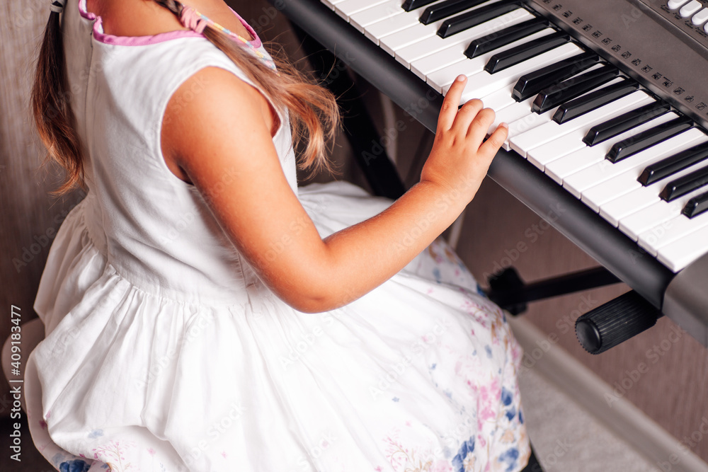 Fototapeta premium Back view a girl in a white summer dress plays an electronic synthesizer in a home interior.