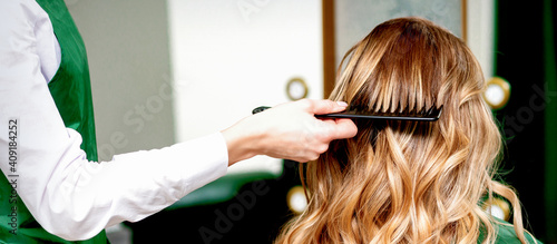 Back view of hairdresser combing wavy hair of a young blonde woman in a beauty salon