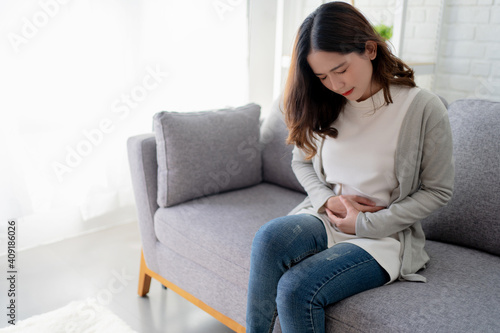 Asian woman has a stomachache sitting on the sofa at home. She feels sick.