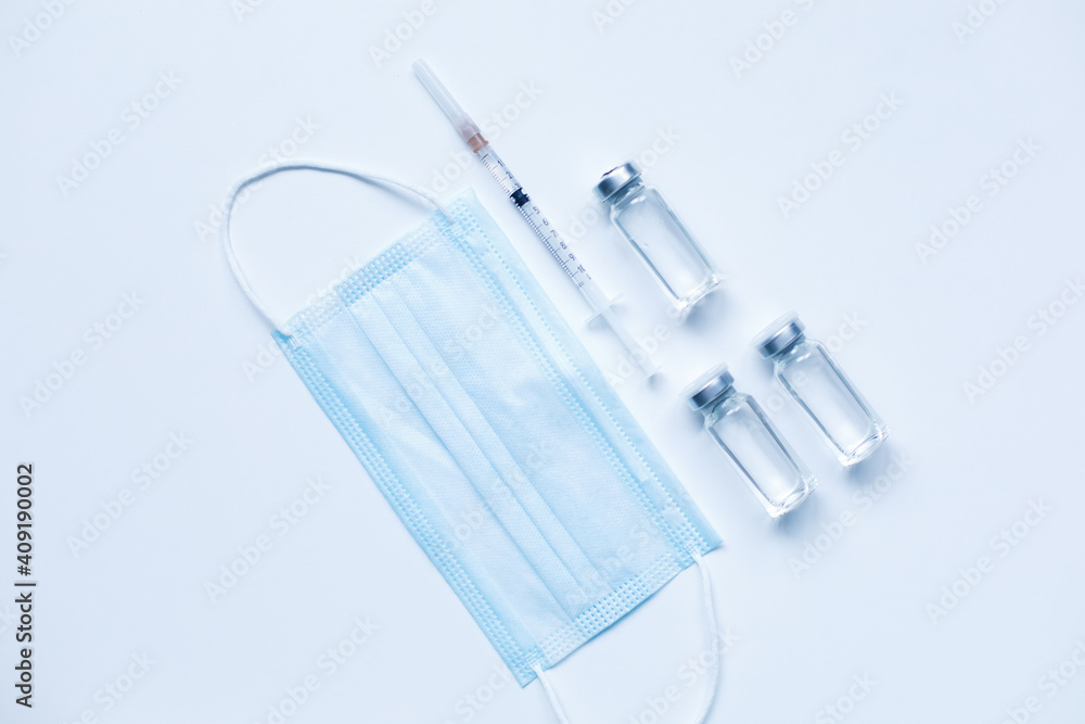 Three glass bottles with liquid , medical mask and syringe on gray background with copy space. The medical conception of combating the covid-19 pandemic. Flu vaccination. Flat lay Three glass bottles with liquid , medical mask and syringe on gray background with copy space. The medical conception of combating the covid-19 pandemic. Flu vaccination. Flat lay