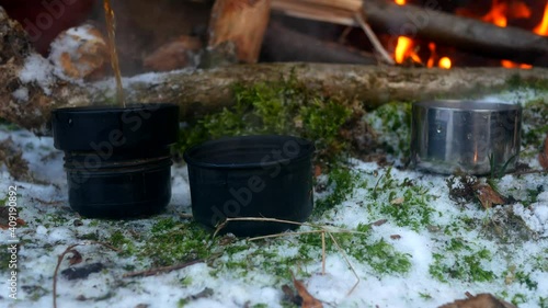 Tea is poured into cups from a thermos near a fire in the winter forest