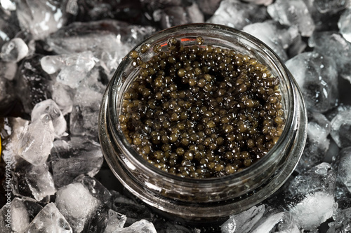 The black caviar of the sturgeon fish in a glass jar is laid on the crushed ice. Top view, copyspace