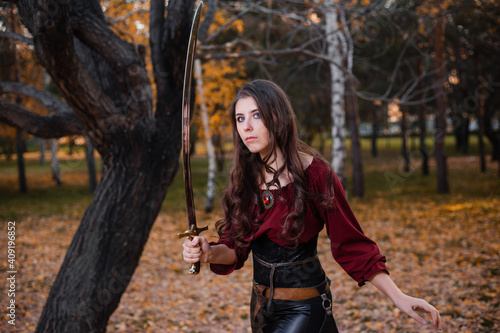 A young woman holds a saber while standing against the backdrop of a gloomy, dark autumn park.