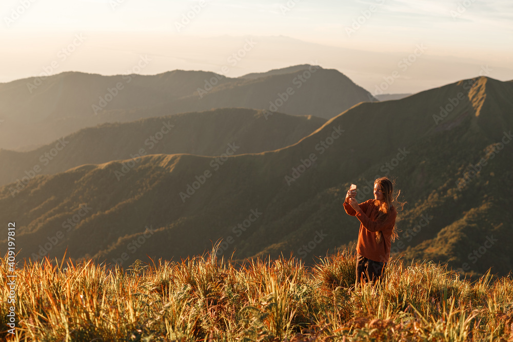 Carefree happy woman enjoying nature on grass meadow on top of mountain with sunrise, taking picture on her phone