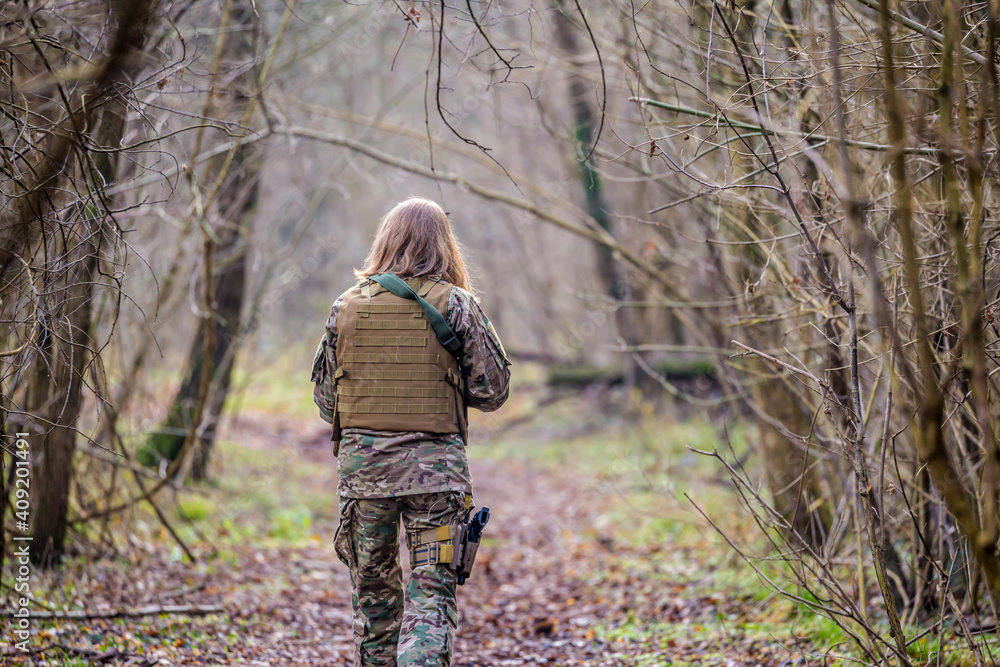 Naklejka premium Girl in military uniform with an airsoft gun walking on a dirt road in the forest