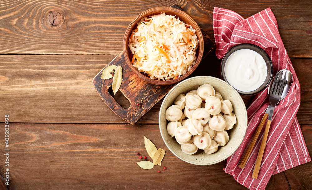 Traditional Russian pelmeni dumplings with sour cream and sauerkraut with sour cream over wooden background. Top view, flat lay. Place for text. Traditional Russian pelmeni dumplings with sour cream and sauerkraut with sour cream over wooden background. Top view, flat lay. Place for text.