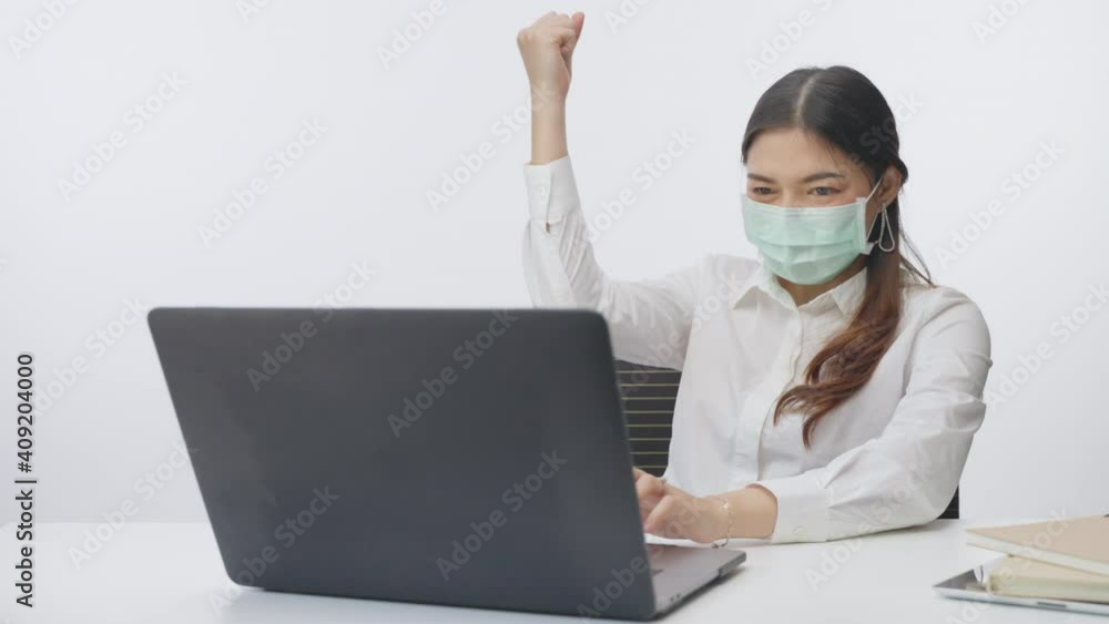 Young happy successful businesswoman wearing surgical mask pump up her hand in excitement in front of her computer during work from home, slow motion with white background Young happy successful businesswoman wearing surgical mask pump up her hand in excitement in front of her computer during work from home, slow motion with white background