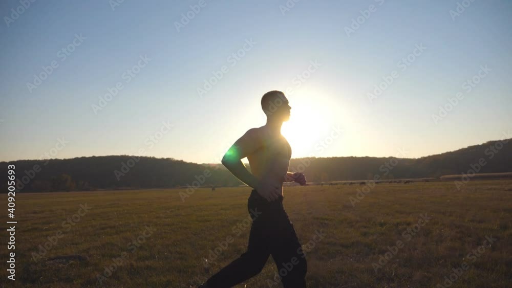 Young muscular man running through field with beautiful landscape at background. Male athlete trains in nature. Soldier jogs at sunset time. Unity with nature. Healthy active rest. Side view Slow mo Young muscular man running through field with beautiful landscape at background. Male athlete trains in nature. Soldier jogs at sunset time. Unity with nature. Healthy active rest. Side view Slow mo