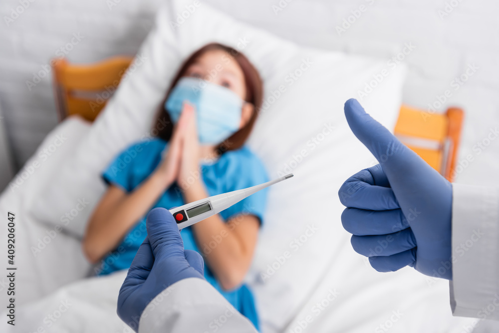 doctor holding thermometer and showing thumb up near child in medical mask lying in bed with praying hands, blurred background doctor holding thermometer and showing thumb up near child in medical mask lying in bed with praying hands, blurred background