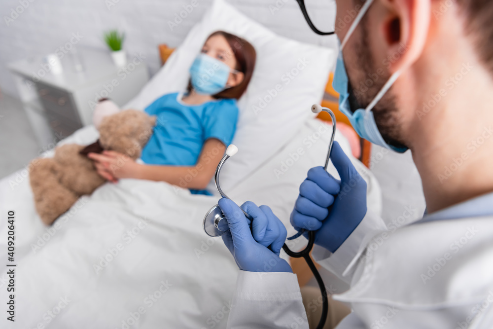 doctor holding stethoscope near sick child in medical mask lying in bed with teddy bear on blurred background doctor holding stethoscope near sick child in medical mask lying in bed with teddy bear on blurred background