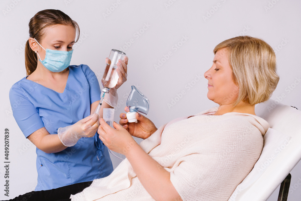 An older patient lies in a chair and a young doctor prepares nebulizer for the treatment of colds and viral respiratory diseases on a white background.
