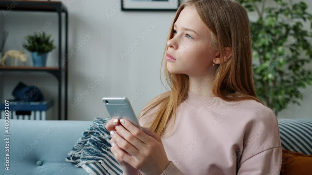 Serious female teenager is using smartphone texting touching screen sitting on couch in apartment alone concentrated on online communication. People and devices concept. Serious female teenager is using smartphone texting touching screen sitting on couch in apartment alone concentrated on online communication. People and devices concept.