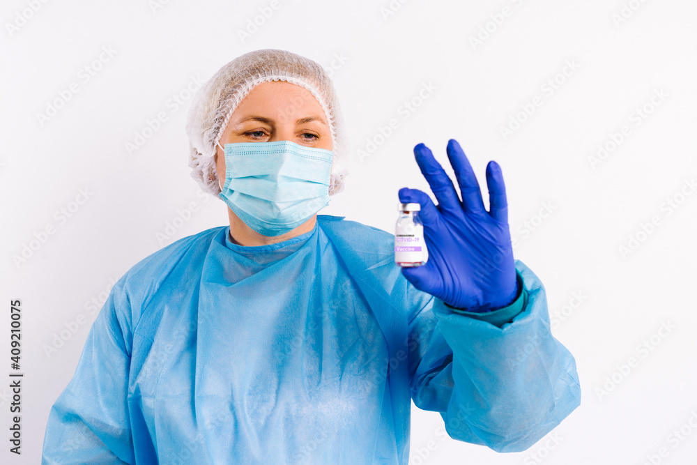 A doctor in protective medical equipment and latex disposable gloves holds an ampoule with a vaccine and shows it to the camera. White background. Fighting covid 19. A doctor in protective medical equipment and latex disposable gloves holds an ampoule with a vaccine and shows it to the camera. White background. Fighting covid 19.