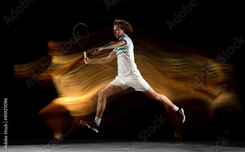 On fire. Young professional sportsman playing tennis isolated on black background in mixed light. Training, practicing in motion, action. Power and energy. Movement, ad, sport, healthy lifestyle