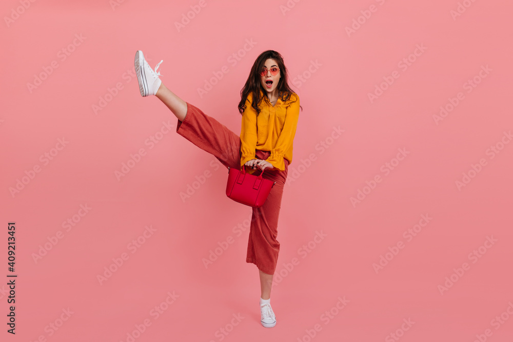 Snapshot of cheerful brunette in pink pants, raising her leg high. Lady in stylish outfit posing with handbag on isolated background Snapshot of cheerful brunette in pink pants, raising her leg high. Lady in stylish outfit posing with handbag on isolated background