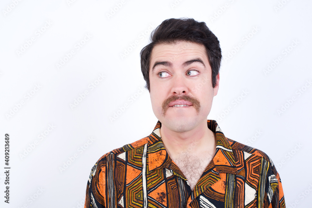 Amazed Young caucasian man wearing generic pattern printed shirt against white wall bitting lip and looking tricky to empty space. Amazed Young caucasian man wearing generic pattern printed shirt against white wall bitting lip and looking tricky to empty space.