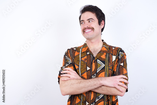 Dreamy rest relaxed Young caucasian man wearing generic pattern printed shirt against white wall crossing arms, looks good copyspace