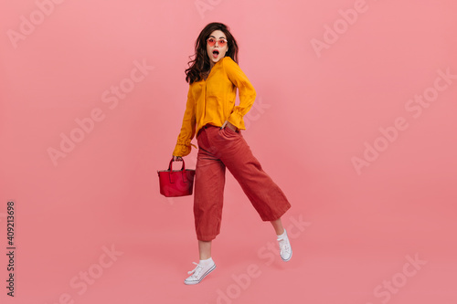 Perky girl in stylish glasses stares into camera in amazement, walking on pink background. Brunette in culottes and orange blouse posing with red handbag photo