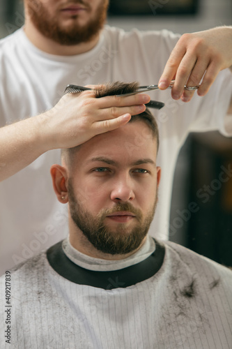 Close up master barber, stylist does the hairstyle to guy, young man. Professional occupation, male beauty concept. Cares of hair, mustache, beard of client. Soft colors and focus, vintage.