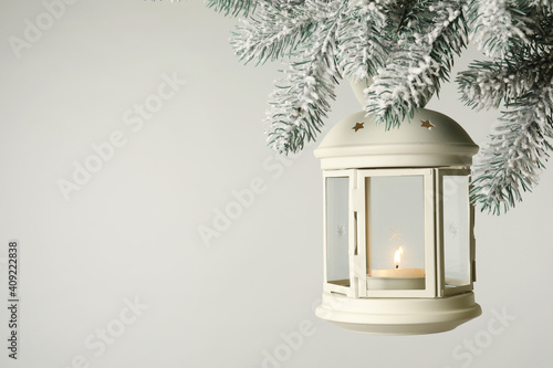 Christmas lantern with candle hanging on snowy fir tree branch against light background. Space for text