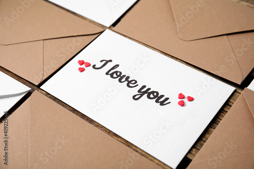 Sheet of paper with phrase I Love You, envelopes and heart shaped sprinkles on wooden table, closeup