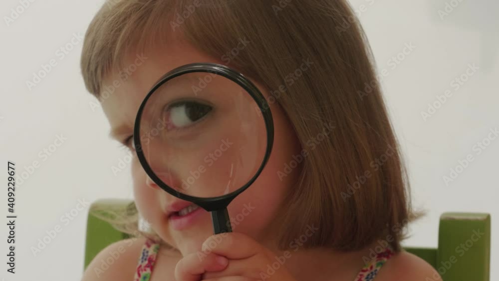 Cute little girl using magnifying glass, talking and smiling, handheld shot. Child looking to camera through magnifier. Kid playing with loupe. Moving lens enlarging eyes, putting it away from face Cute little girl using magnifying glass, talking and smiling, handheld shot. Child looking to camera through magnifier. Kid playing with loupe. Moving lens enlarging eyes, putting it away from face